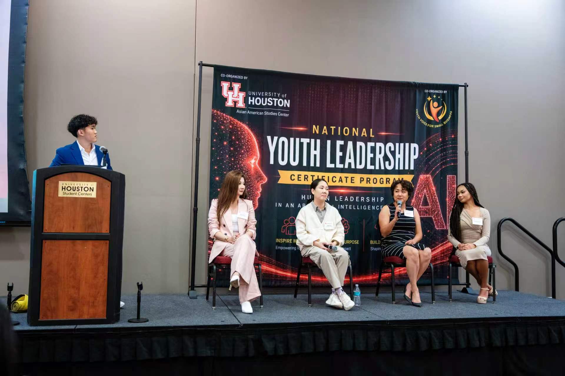 Student moderator Simon Zhong stands at the podium with four panelists sitting on stage to answer questions.