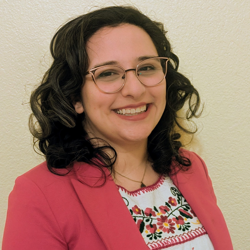 A curly-haired woman smiles while wearing gold glasses, a pink blazer, and oaxacan shirt.