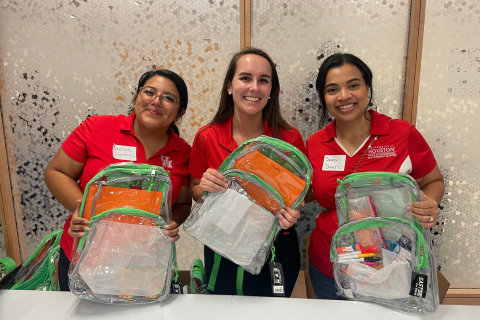 Three women stand side by side smiling and holding up clear backpacks filled with school supplies, including notebooks, folders, and pencil cases. They are wearing matching red polo shirts with University of Houston logos and name tags, indicating they are volunteers. The background features a textured, frosted glass wall. The atmosphere appears cheerful and welcoming, suggesting participation in a community back-to-school event.