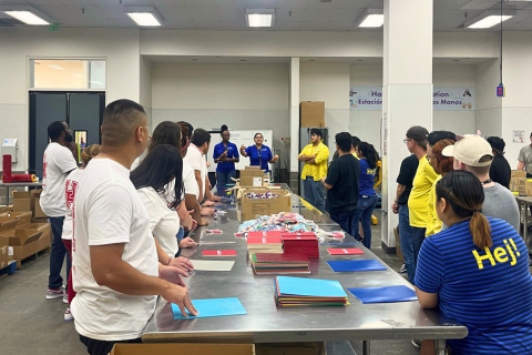 A large group of volunteers stands around a long metal table in a warehouse-like setting, preparing for an organized activity. The table is covered with stacks of colorful paper, string-bound packets, and other materials. Most volunteers wear casual clothing, with some in matching yellow or white shirts. At the far end of the room, two people in blue shirts appear to be giving instructions to the group. The room is brightly lit with fluorescent lights, and boxes and supplies are stacked along the walls. The environment suggests a community service or group assembly event.