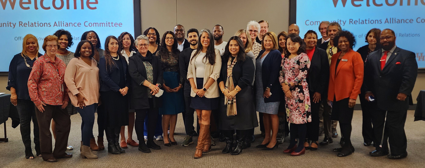 Group photo of the Community Relations Alliance members standing together in a conference room, with a large title reading Community Relations Alliance Committee