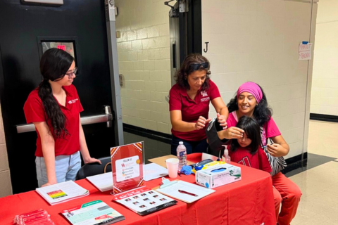 Three women and one young girl are participating in an activity at a school hallway event. Two women in red shirts, representing the University of Houston, assist a seated woman with a pink headband who is smiling while helping a young girl. The girl is seated and receiving a vision screening or similar health check. A red table in front of them is covered with informational flyers, a sign-up sheet, medical supplies, and a box of masks. The setting appears to be a community outreach or back-to-school health fair held indoors near classroom doors.