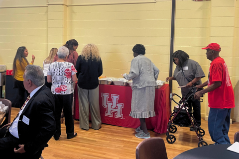 A group of people stands in line at a buffet table draped with a red University of Houston tablecloth, selecting food items. The event takes place in a community center or cafeteria with yellow walls and wooden floors. Attendees include a mix of older adults, one using a walker, and others in casual or semi-formal attire. A few people are seated at round tables in the foreground. The setting suggests a casual, university-sponsored community gathering or meal event, possibly aimed at outreach or engagement.