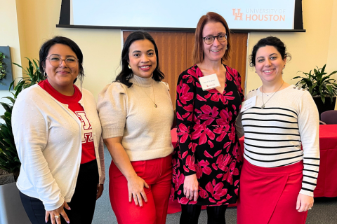 Four women stand together smiling in a brightly lit room with plants and a projection screen in the background displaying the University of Houston logo. Three of the women wear red and white outfits, while the fourth, who stands in the center, wears a black dress with a bold red floral pattern and a name tag. The setting appears to be a professional or academic event, likely a symposium or conference focused on community engagement and experiential learning. The atmosphere is friendly and professional.