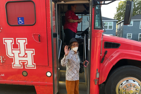 An elderly person wearing a mask, glasses, a black hat, and patterned shirt waves while stepping off a bright red University of Houston bus. The side of the bus features a large "UH" logo and a wheelchair accessibility sign. A woman in a red shirt stands inside the bus near the driver’s seat. The setting appears to be a neighborhood street lined with houses. The moment captures a cheerful and community-focused initiative, likely involving transportation support for civic engagement such as voting.