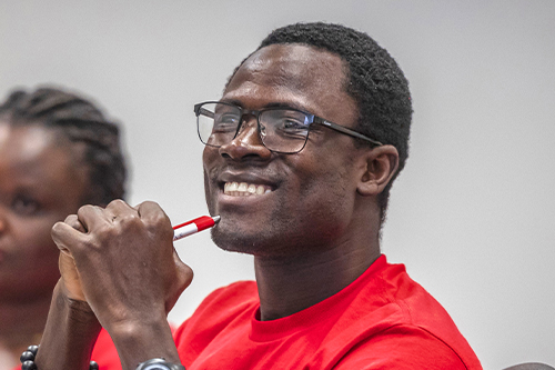 Black male student smiling with a red shirt clasping a pen in class
