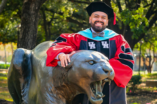 A man standing with his arm around a statue of a cougar - he is in red and blue graduation regalia with trees in the background