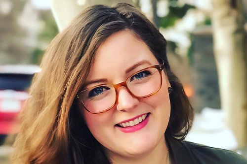 A white woman smiling with light brown hair, glasses with trees in the backgroud