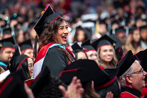 A female student with black hair in graduation regalia smiling at a commencement ceremony