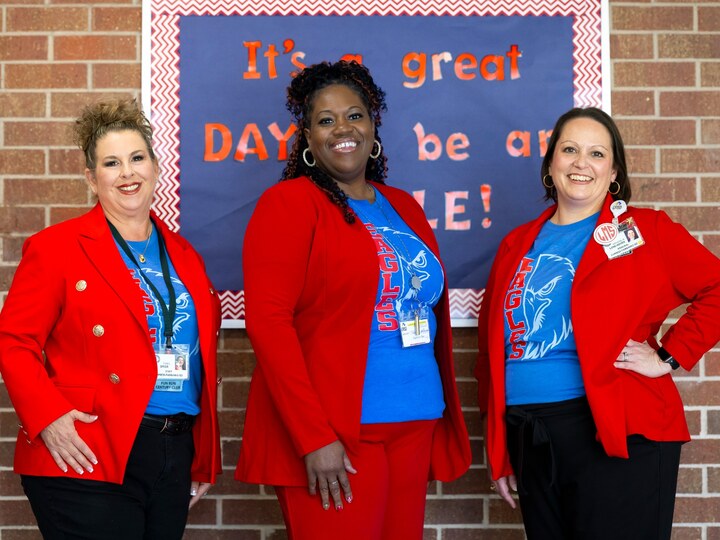 Three school leaders pose in front of a brick wall and school sign.