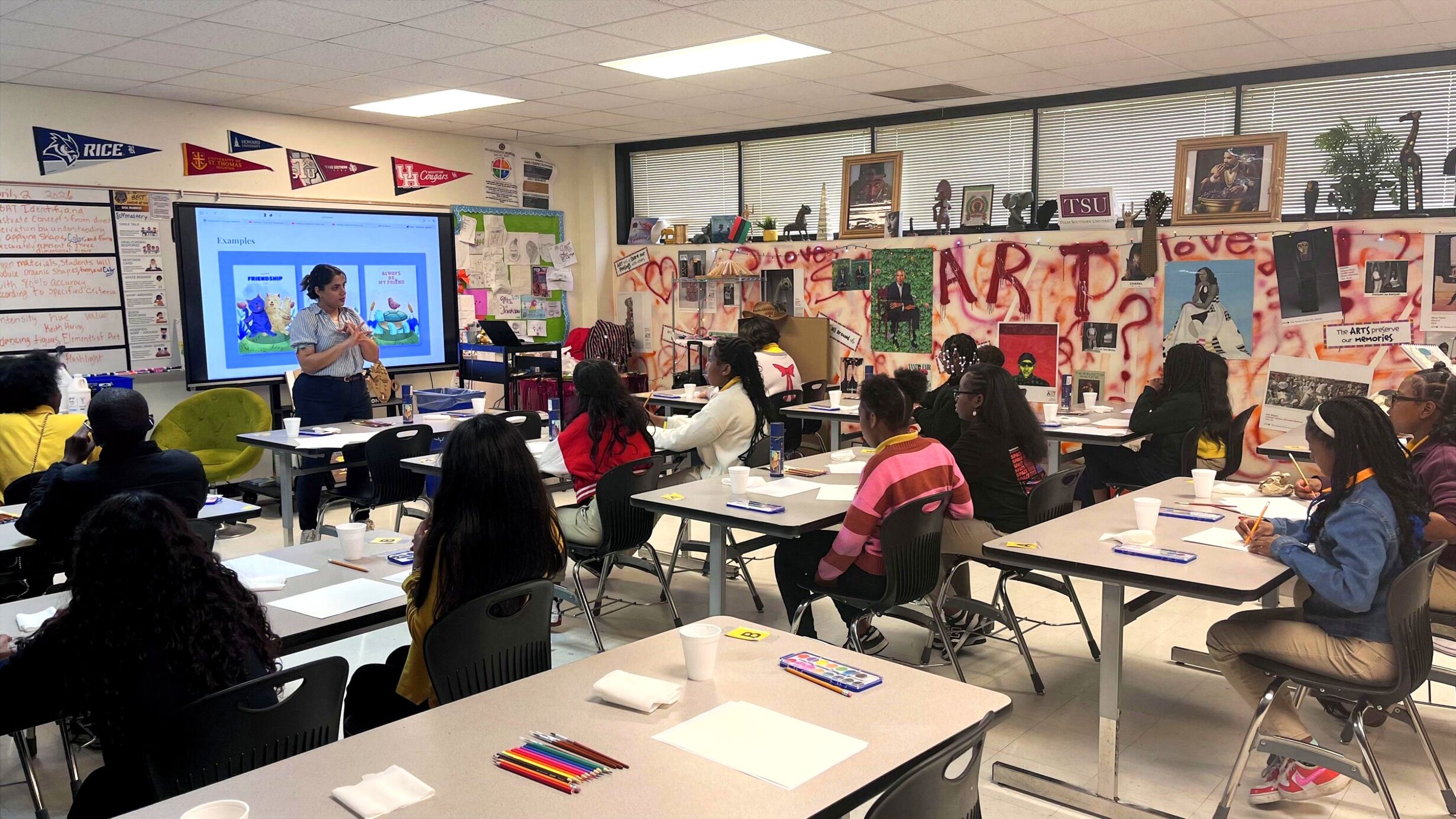 A classroom with students seated at tables, listening to a teacher near a large screen. The room is decorated with colorful art and college pennants, conveying a creative, educational atmosphere.