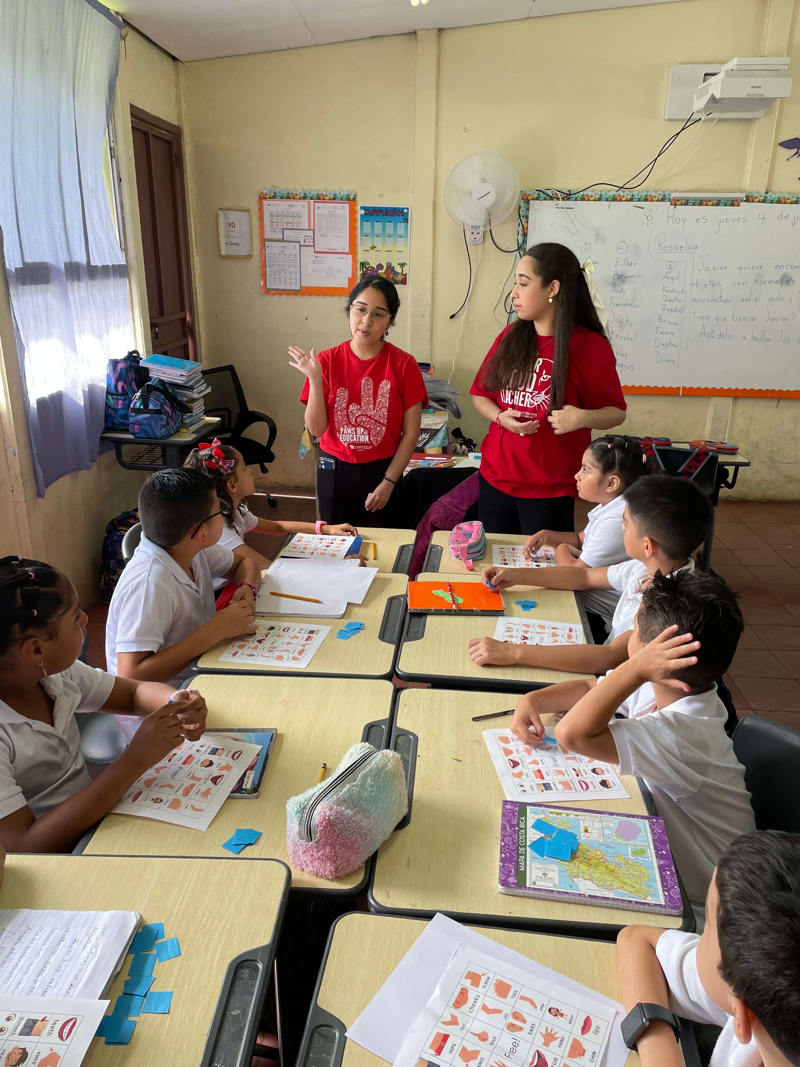 Two UH students teach a group of students with picture worksheets at their desks.