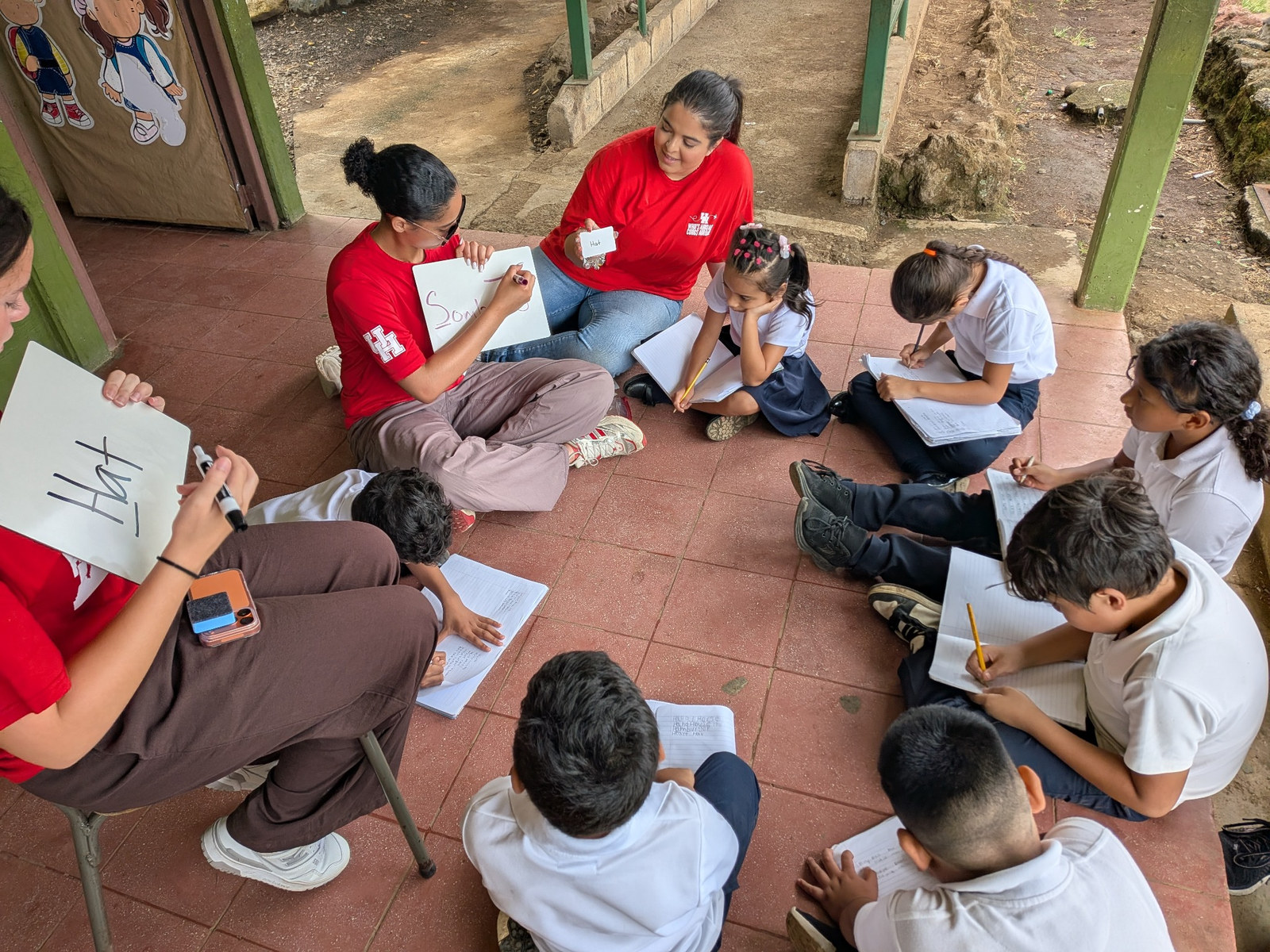 Three student teachers use whiteboards and flashcards to teach Costa Rican children sitting outside in a circle.