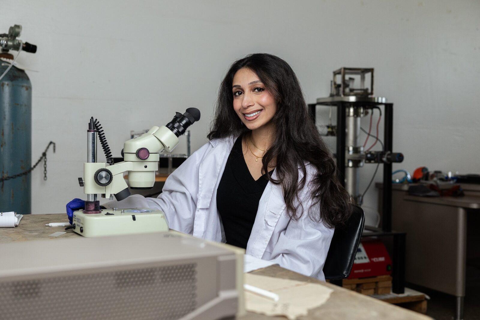 Salena Kabani wears a lab coat and sits in front of a microscope in a research lab.