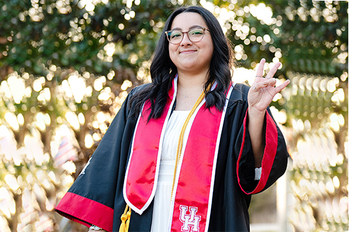 A female college student in regalia with brown hair and glasses flashing the Coog sign outside among the trees
