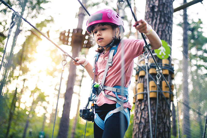 Young girl on a zipline with a pink shirt and helmet. She is in the woods.