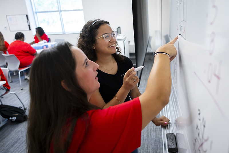 Two college students working at a white board; they both have brown hair; one has a red shirt; one has a black shirt and glasses