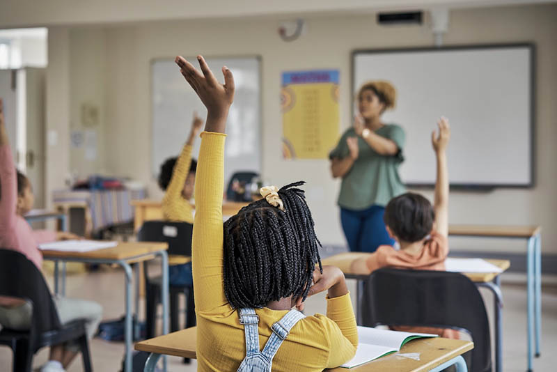 A black female teacher in front of a class of students with their hands raised