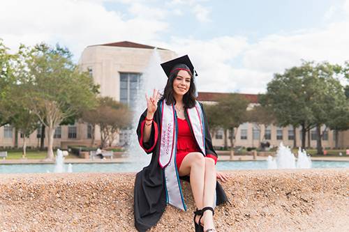 Female student in regalia sitting in front of a UH building and founntains
