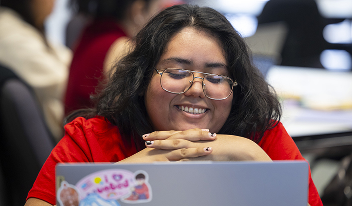 Student with brown hair and glasses, smiling and looking at a laptop