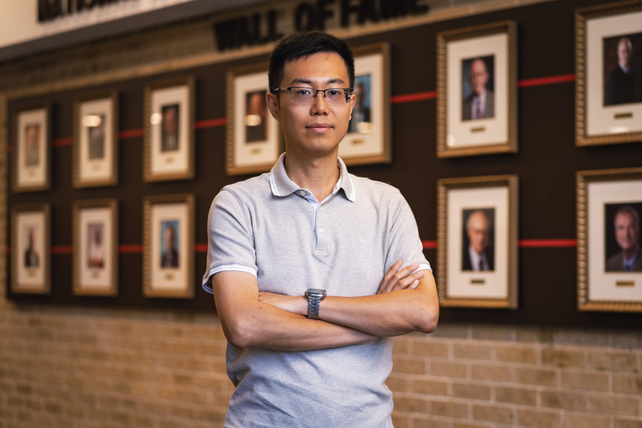 Zheng Fan, wearing a light-colored polo shirt, stands with his arms crossed in front of a dark wall with rows of portraits.