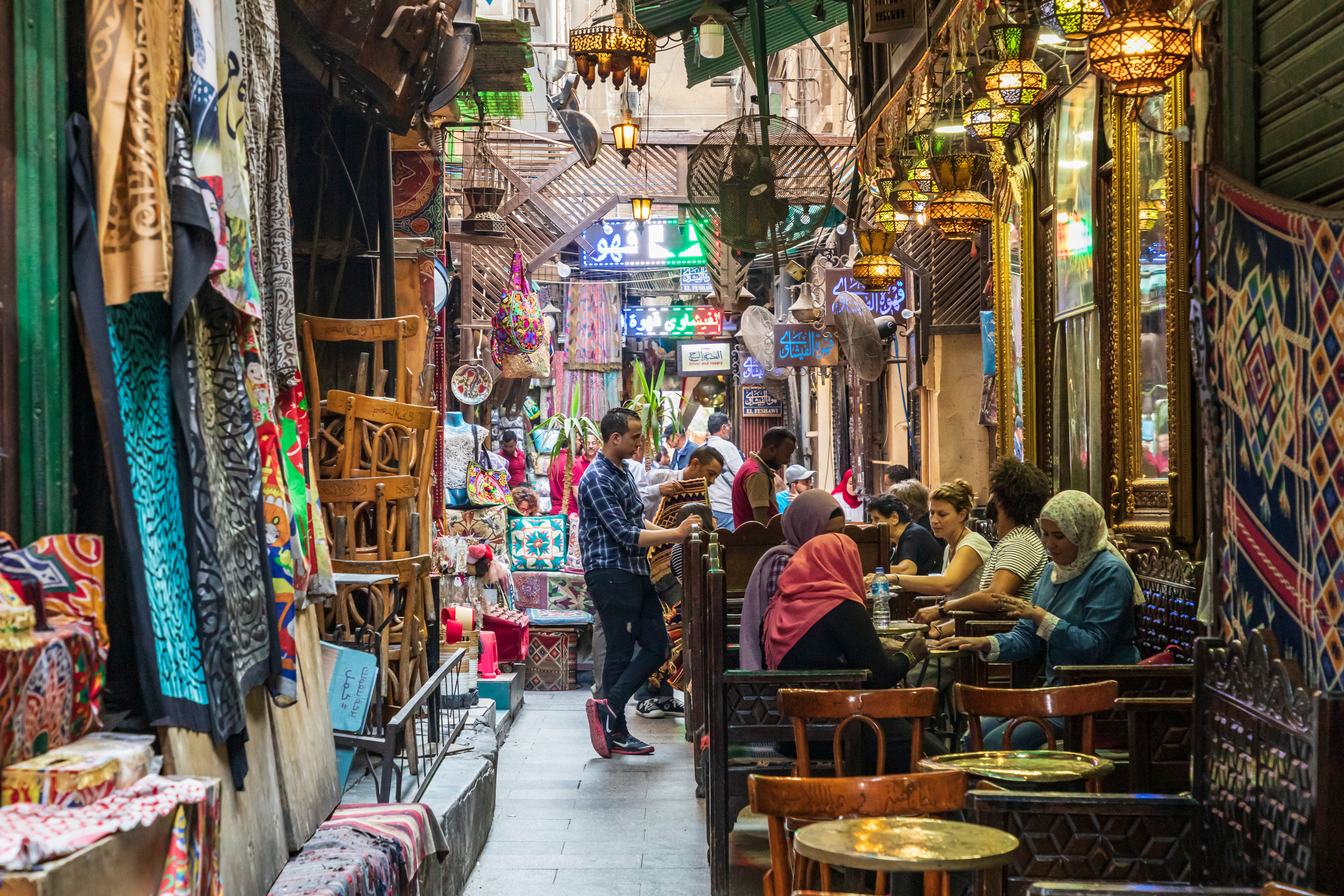 A vibrant, narrow street market with people dining at outdoor tables and colorful textiles, lanterns, and crafts on display.