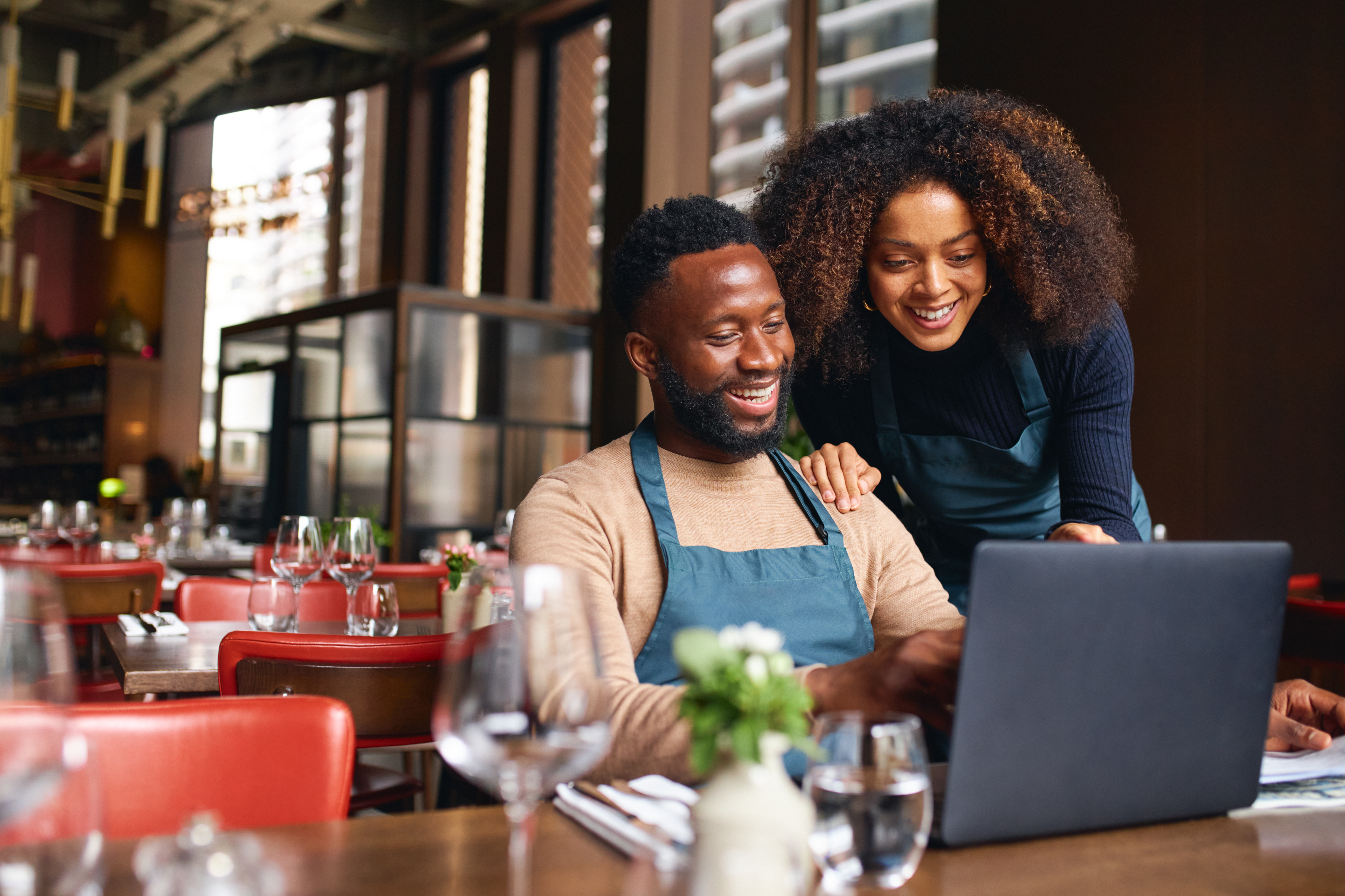 Two restaurant workers smiling while looking at a laptop in a modern dining space.