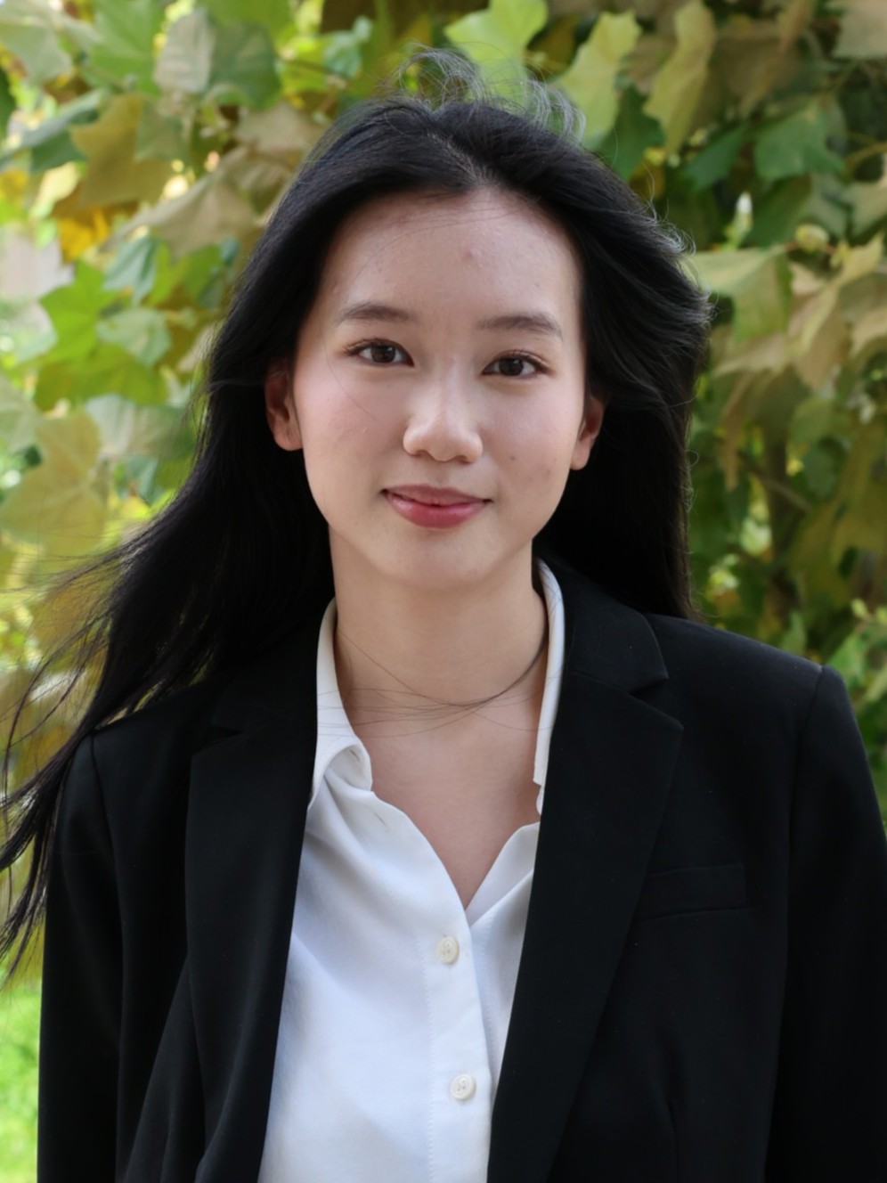 Professional outdoor portrait of Han Nguyen looking at the camera, wearing a black blazer and white blouse against a softly focused leafy background.