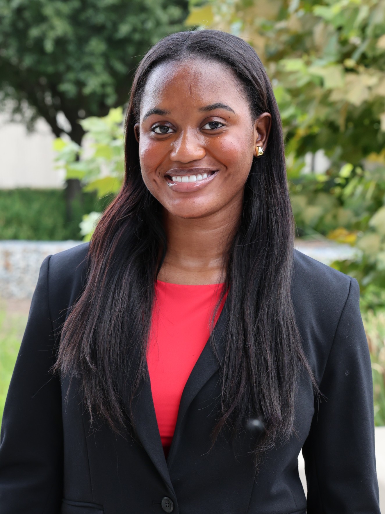 Professional outdoor portrait of Jamila Sankoh smiling at the camera, wearing a black blazer over a bright red top, with leafy greenery in the background.