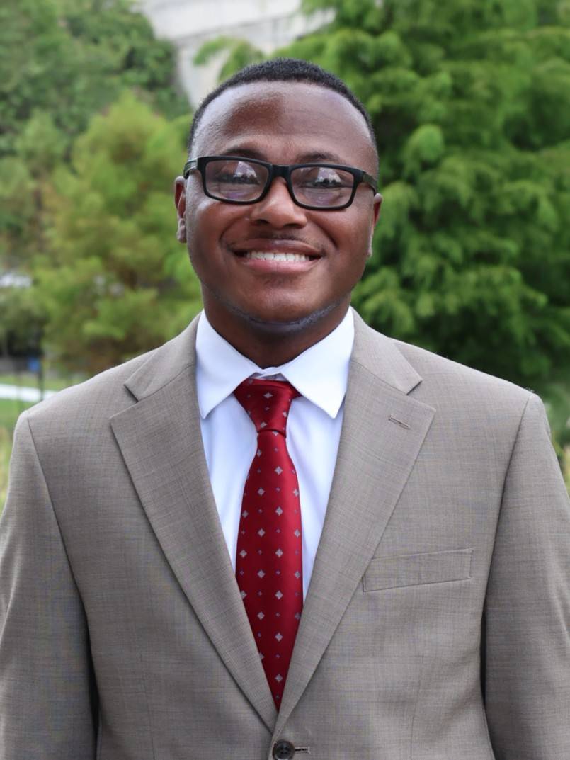 Professional outdoor portrait of Joshua Barlow smiling at the camera in black glasses, a light gray suit, white shirt, and patterned red tie, with trees behind him.