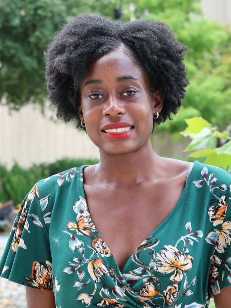 Professional outdoor portrait of Maurya Rodrigues smiling gently at the camera, wearing a green floral dress and gold hoop earrings, with greenery in the background.