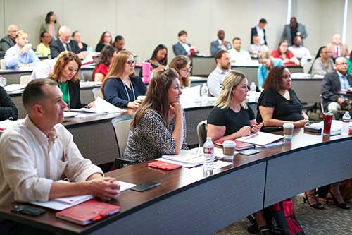 A group of professionals sit in curved rows of desks in a lecture hall.