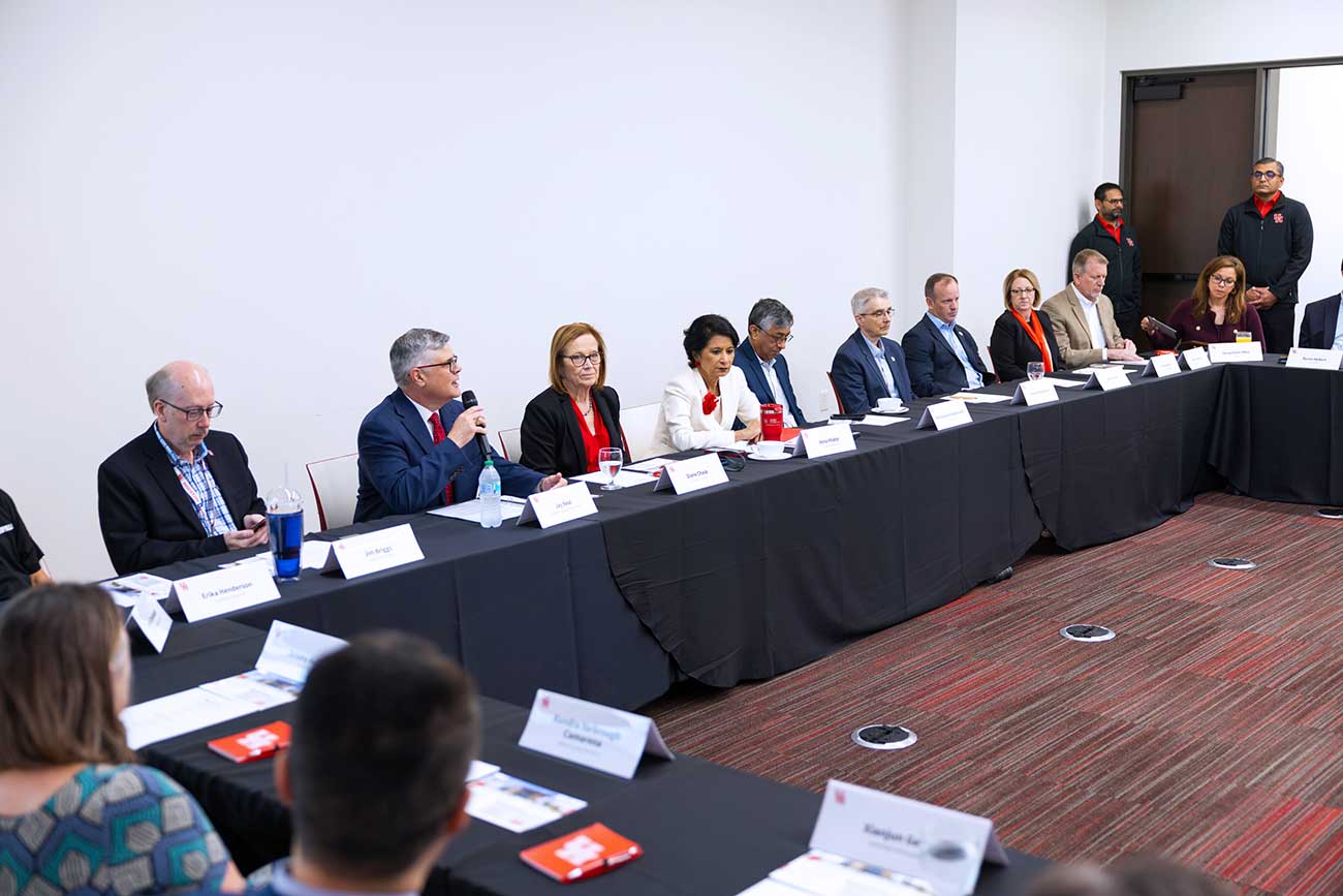 A group of men and women in business suits sit along row of tables with black cloths. A man with glasses in the middle speakins into a microphone.