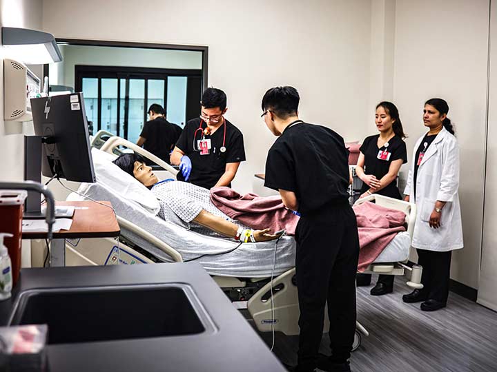 A two nursing students flank hospital bed with a high-fidelity manikin while another student and an instructor watch.