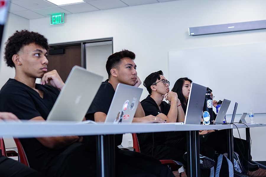 A group of students in scrubs sit at a desk with laptops listening to an out-of-frame instructor.