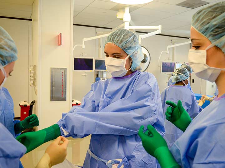 Nursing students wearing surgical masks and hair covers put on gowns and gloves.