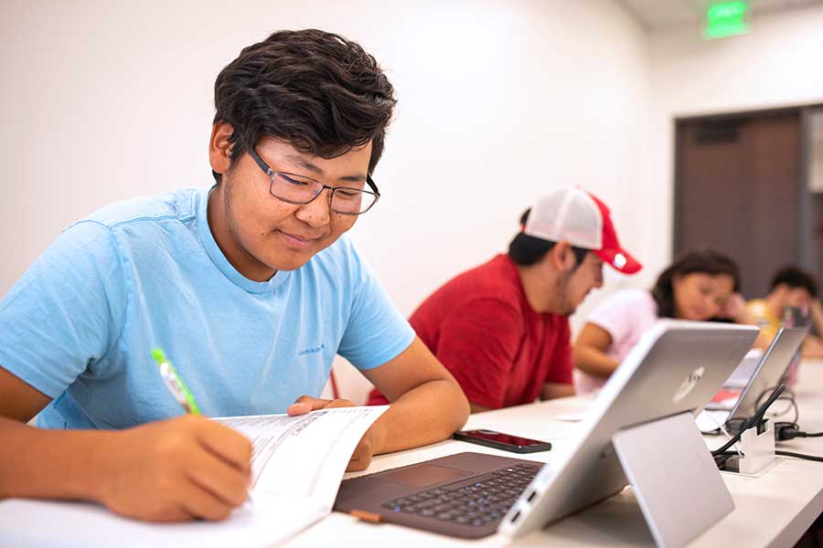 Close up of students sitting at a row in a classroom. The students are writing on papers and looking at laptops.