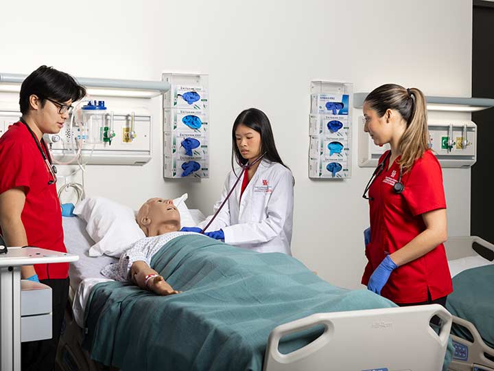A nursing student uses a stethoscope on a high-fidelity manikin in hospital bed while two students watch.