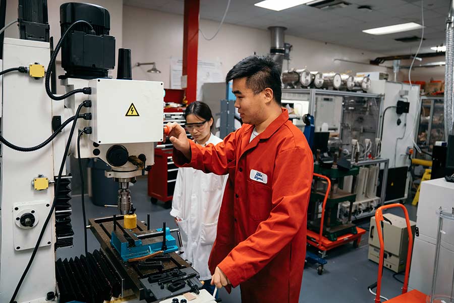 A man and a woman in jumpsuits work in a room with several large pieces of equipment. 