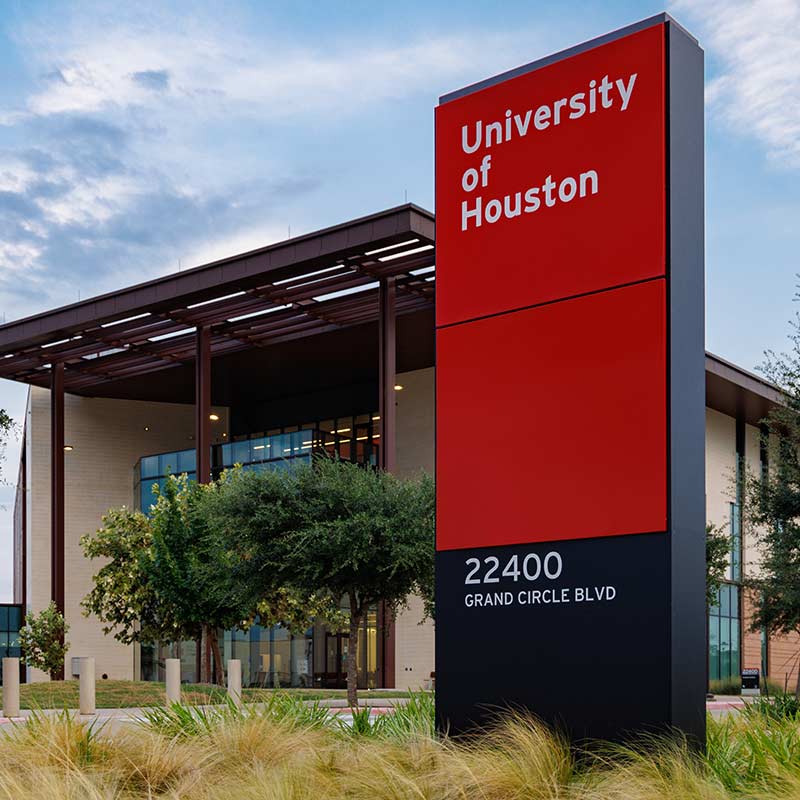 Photograph of a tall red monolith sign with the words 'University of Houston'. A three story birck and metal building is in the background.