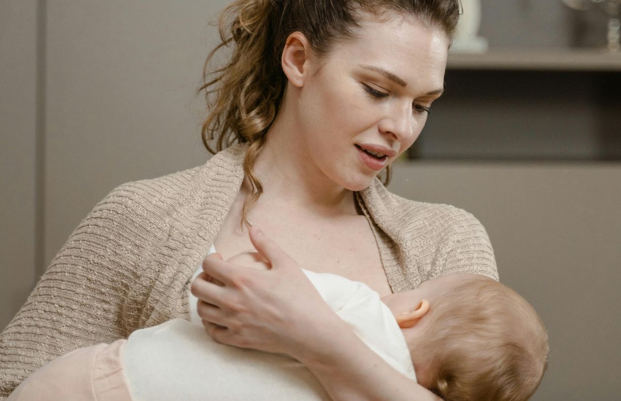 Woman breastfeeds a baby while looking down with a gentle expression in a home setting.
