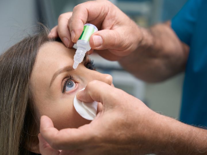 Person receives eye drops from a healthcare professional, with a cotton pad held under the lower eyelid.