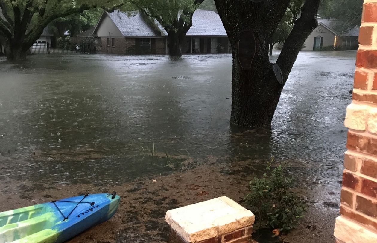 Floodwaters cover a residential street and yards, reaching tree trunks and houses, with a kayak resting near the porch.
