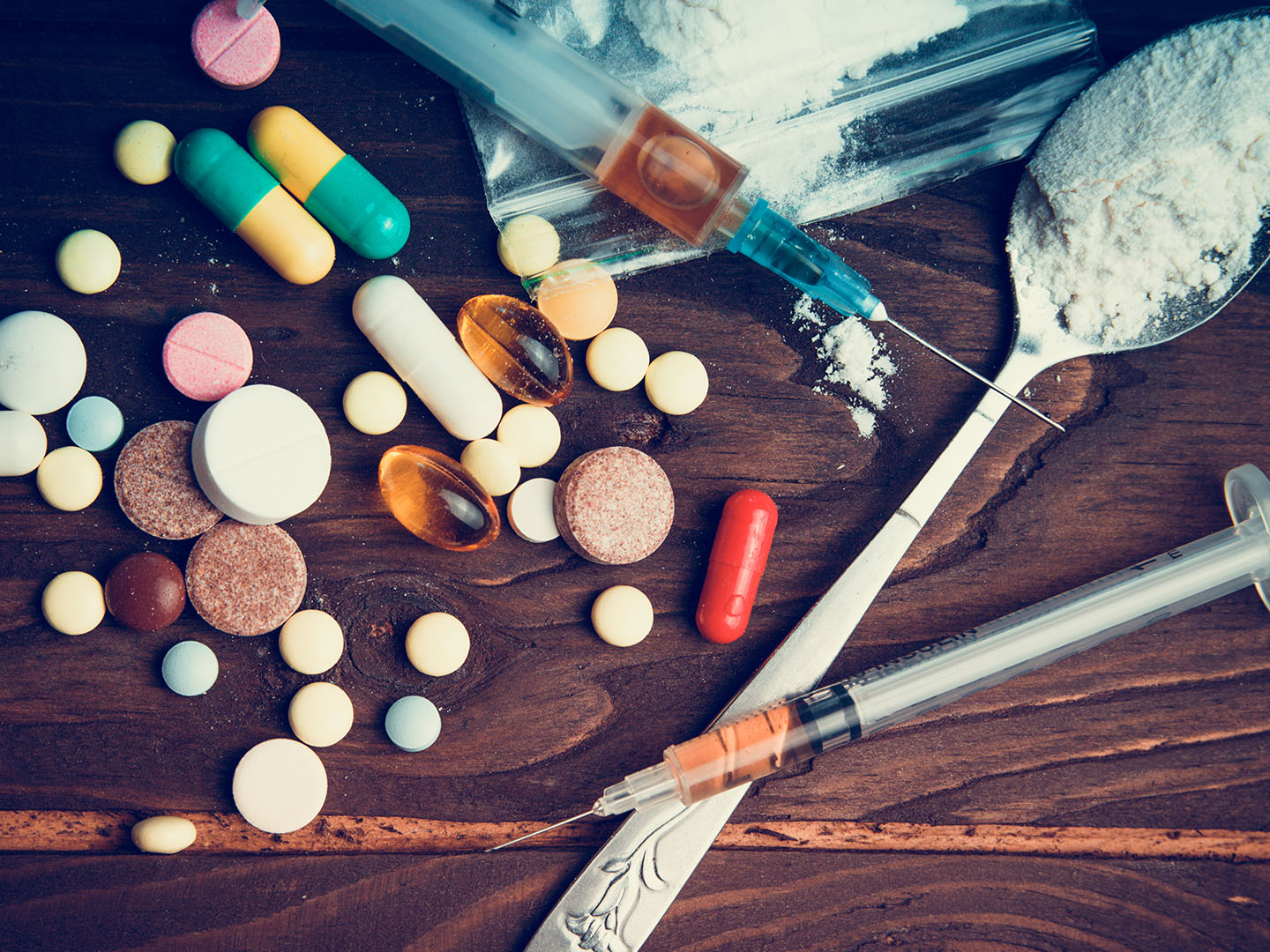 A variety of pills, capsules, and tablets scattered across a wooden surface alongside drug paraphernalia, including syringes, a spoon with white powder and a small plastic bag containing powder.