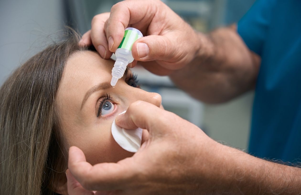 Person receives eye drops from a healthcare professional, with a cotton pad held under the lower eyelid.