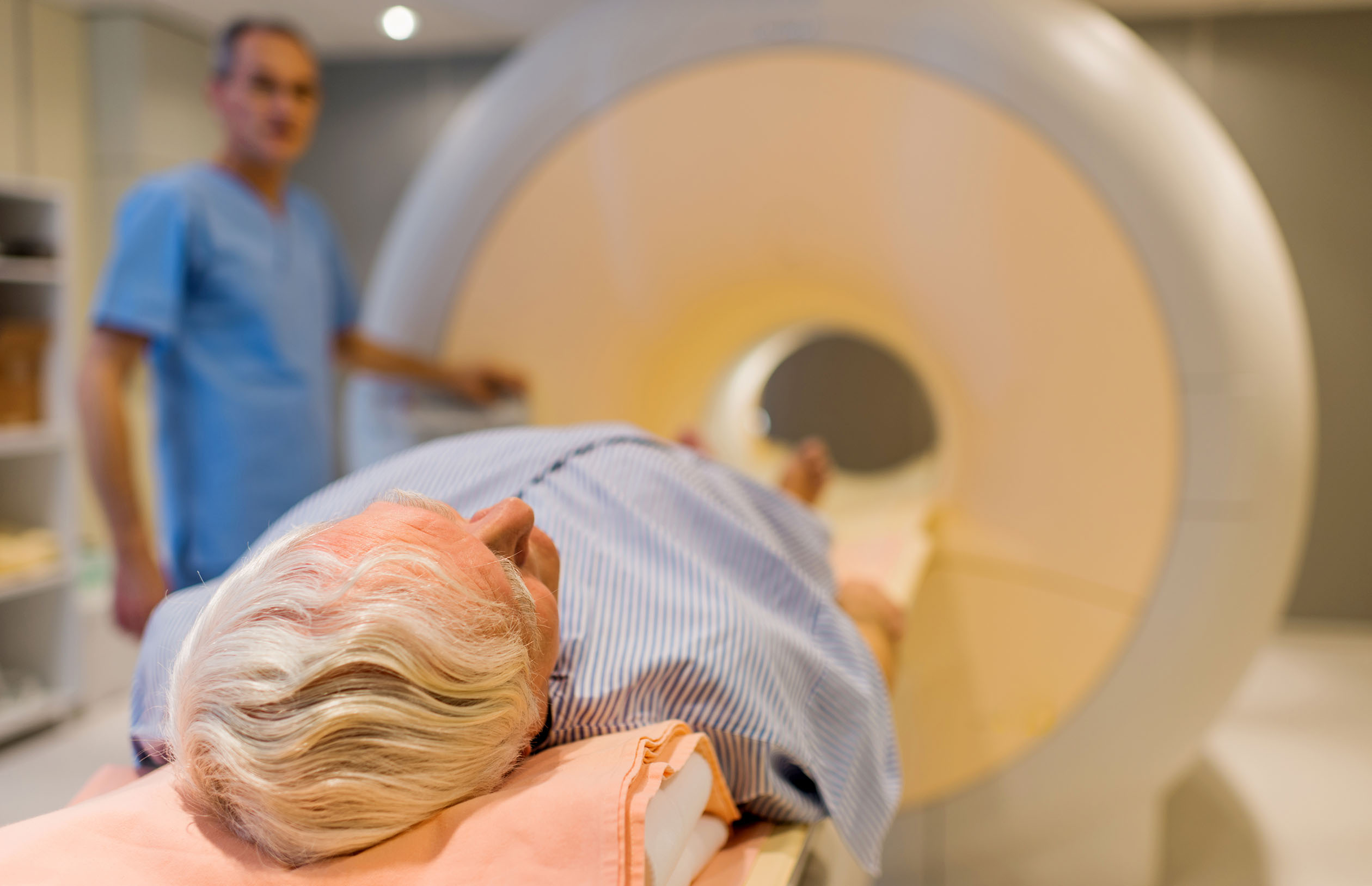 Senior male patient lying down at MRI scan. Radiologist is in the background.