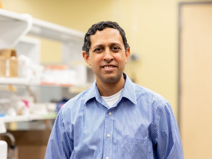 Navin Varadarajan smiling at the camera in a laboratory setting, wearing a striped shirt.