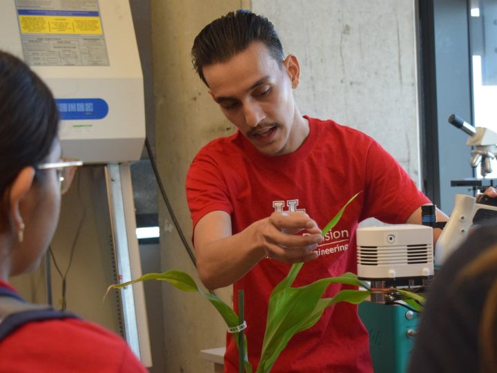 Person in a red University of Houston shirt conducting a scientific demonstration with plants and laboratory equipment. Person in a red University of Houston shirt conducting a scientific demonstration with plants and laboratory equipment.