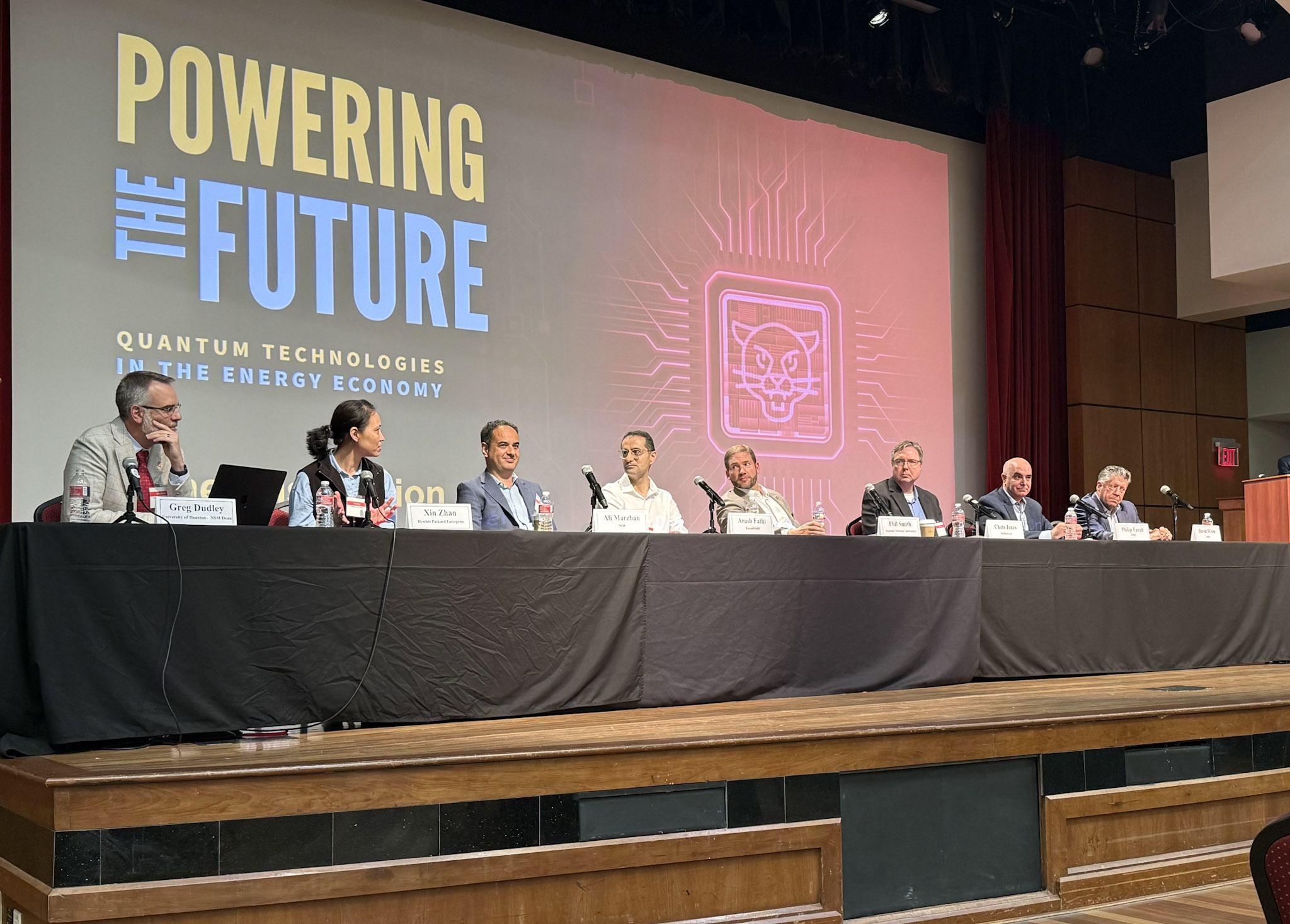 Panel of people in suits sitting at a table with microphones in front of a projection screen.