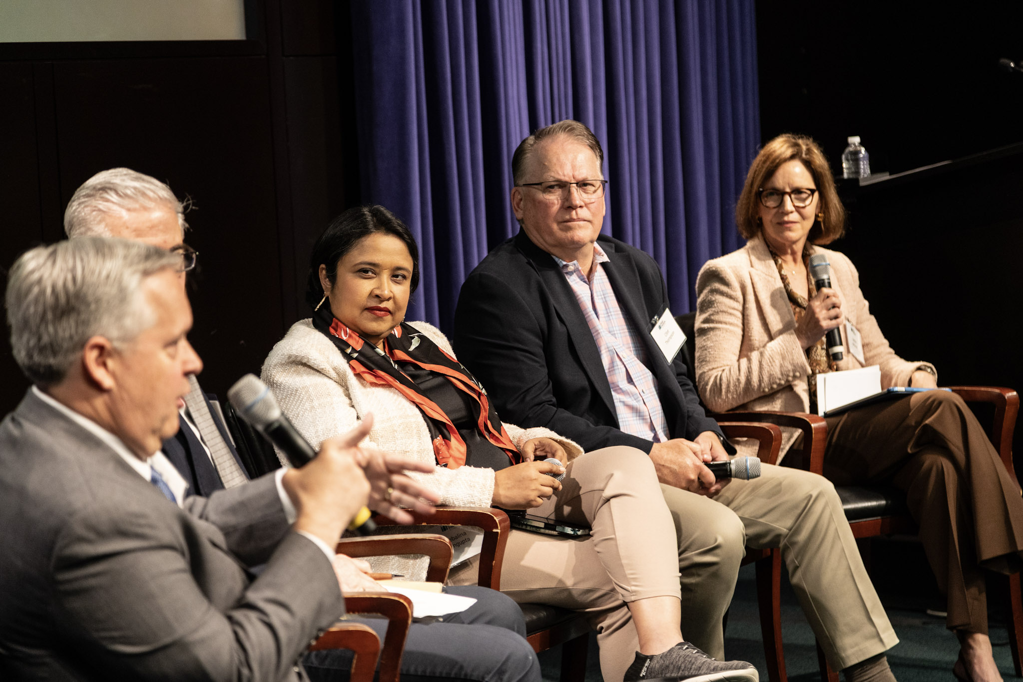 Five people in business attire sitting on stage with microphones, engaged in a panel discussion.